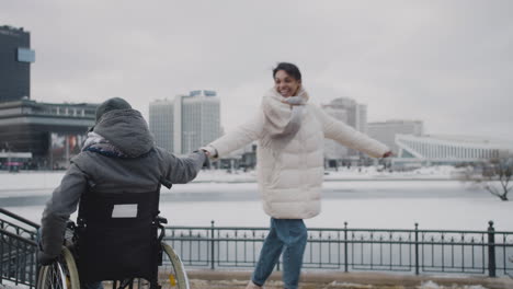 mujer musulmana feliz y su amiga discapacitada en silla de ruedas girando tomados de la mano en la ciudad en invierno 1