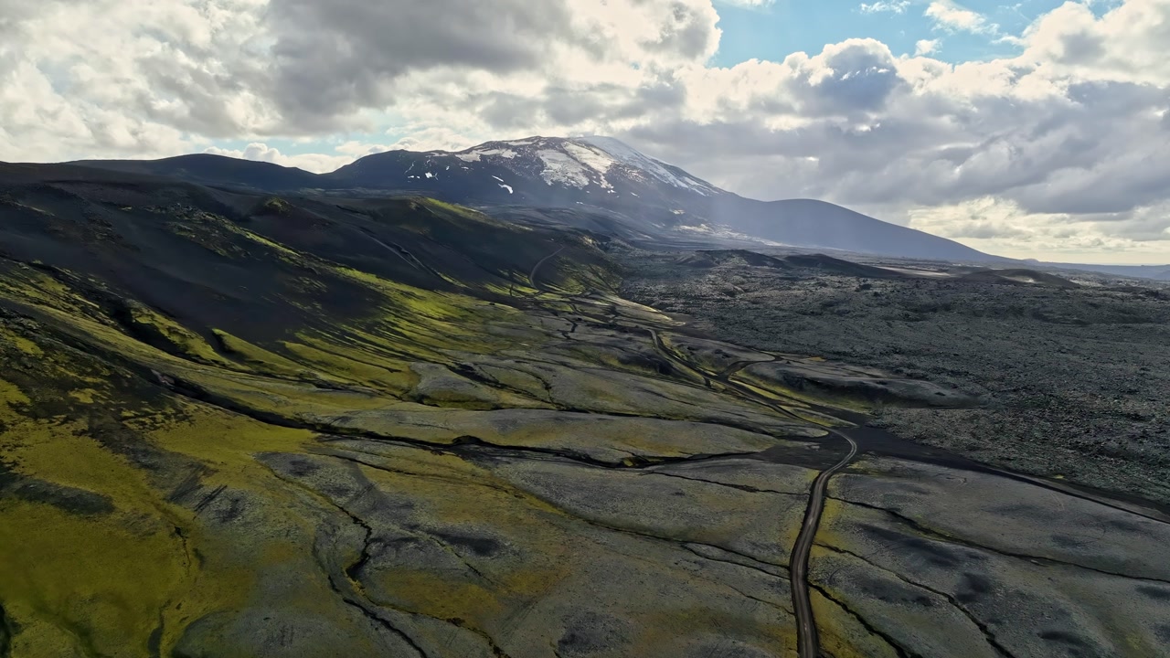 Aerial view of a winding road crossing mossy lava flats toward a snow-capped volcano, with dramatic clouds and sun rays over a vast remote highland landscape.