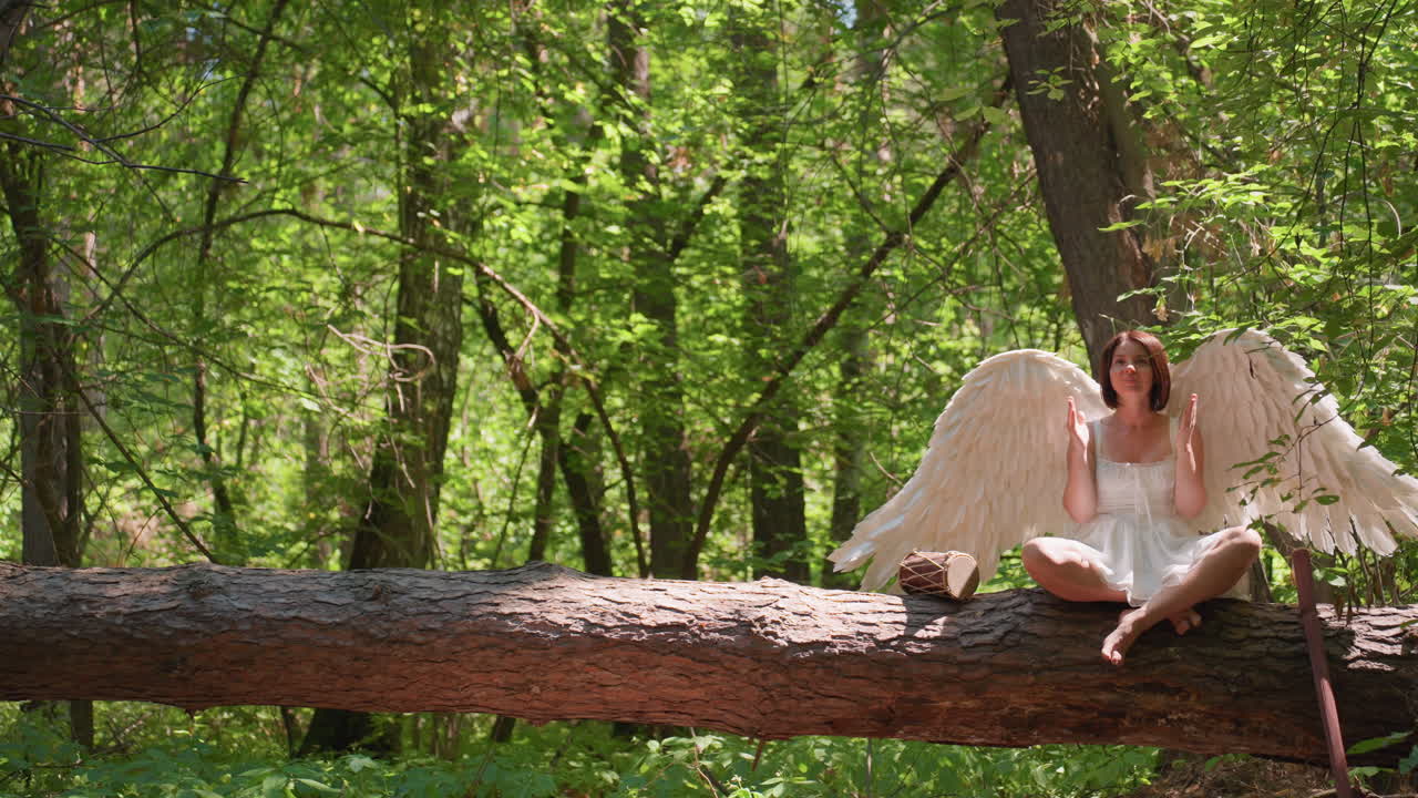 Forest guardian with angelic wings dressed in white sits cross-legged on fallen log praying to gods beside talking drum and wooden staff under gentle sunlight surrounded by green leaves