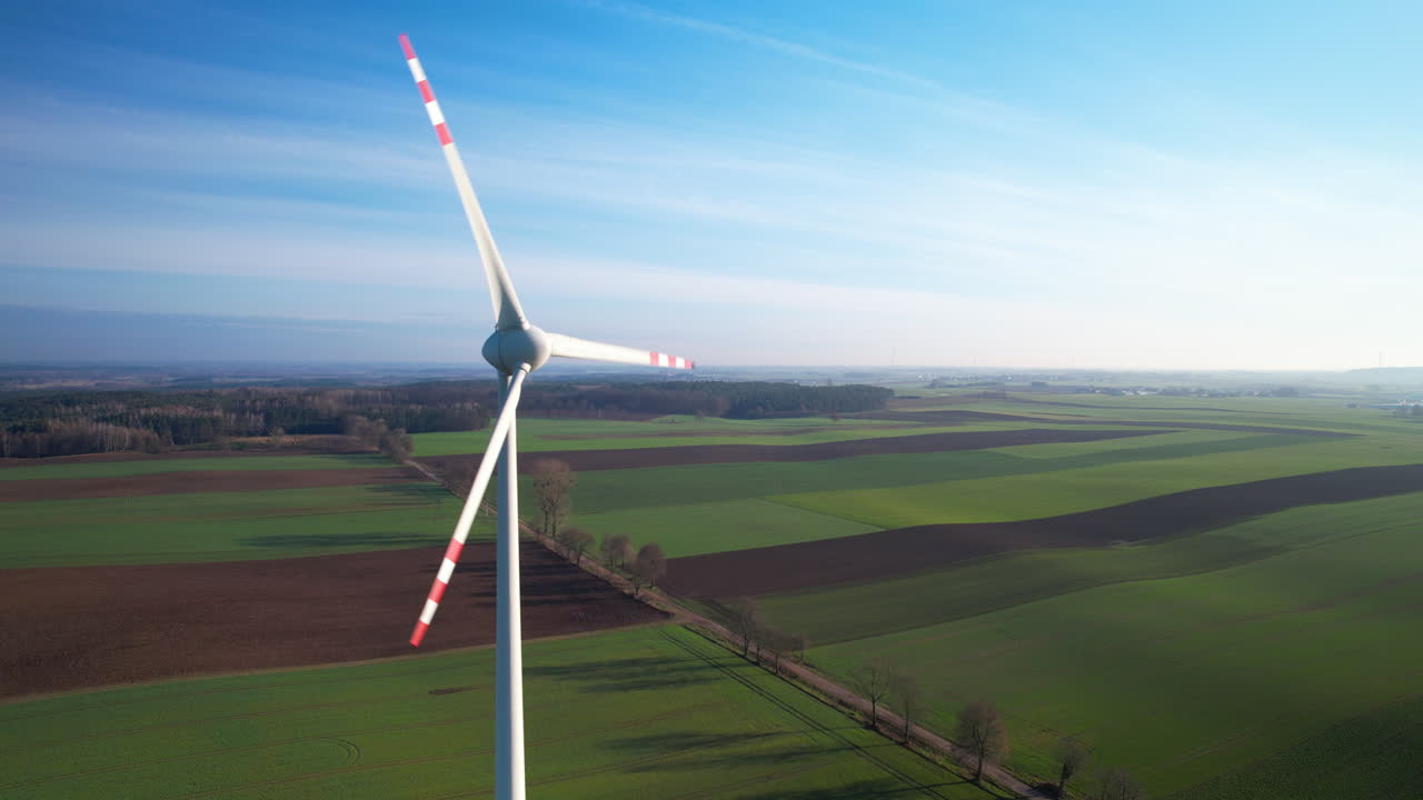Ripresa con drone di una turbina eolica in rotazione nella campagna rurale polacca, sullo sfondo di campi agricoli verdi e una strada di campagna al tramonto - allontanamento.