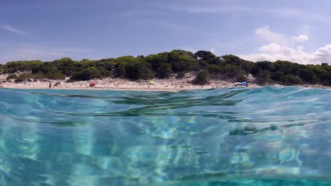 Half-underwater-scene-of-little-red-haired-girl-swimming-beneath-waterline-surface-in-incredible-crystal-clear-tropical-sea-water-of-Saleccia-beach-in-Corsica-island,-France