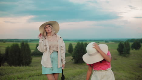 woman in sun hat holding it with a pose while daughter in pink adjusts her own hat, both stand against a scenic countryside with rolling green hills under a vast blue sky