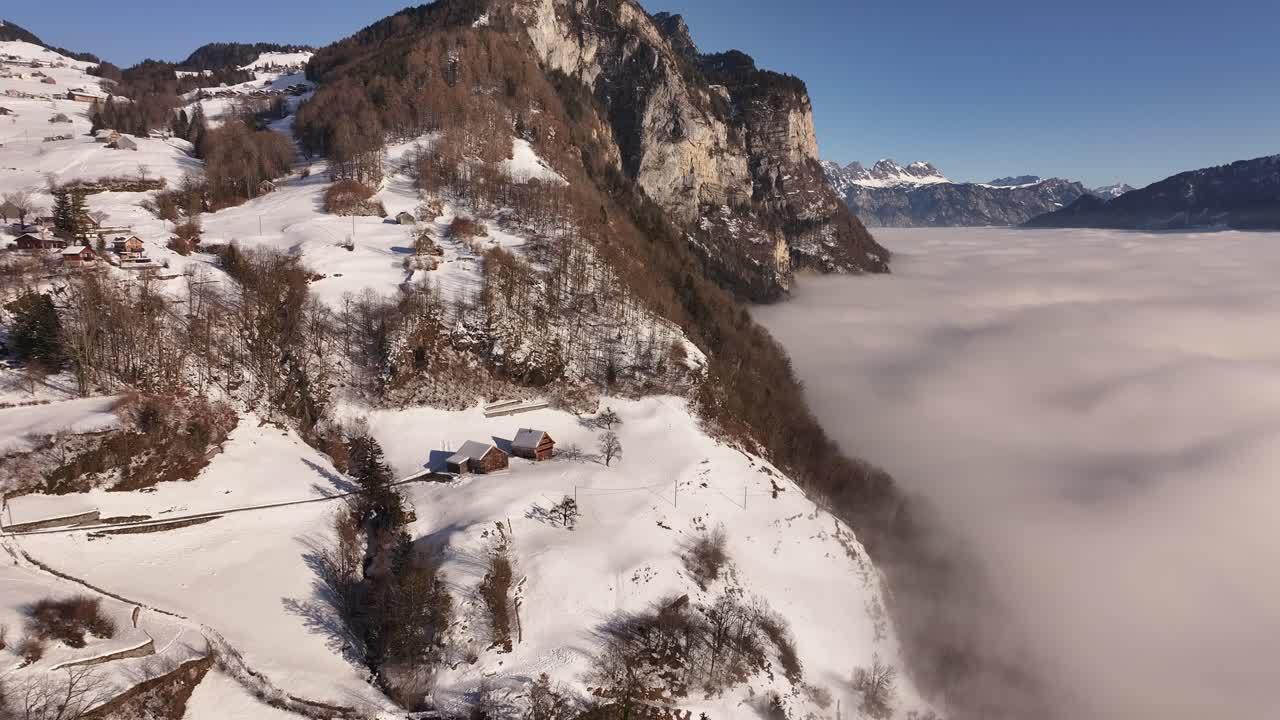 Aerial view of a winding road cutting through a snowy winter landscape in Amden, Switzerland, with a towering cliff face rising above a thick sea of fog, creating a dramatic and serene scene.