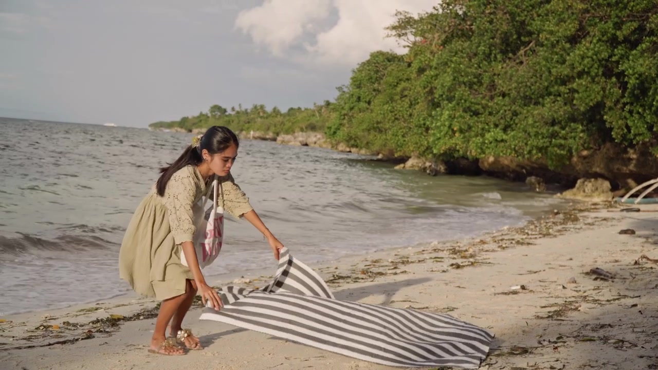 Donna asiatica stende un asciugamano a righe sulla spiaggia. A piedi nudi in un vestito floreale dispiega una coperta sulla sabbia vicino a una costa di mangrovie verdi, lisciando il tessuto mentre dolci onde lambiscono la riva, luce dorata calda, soffusa