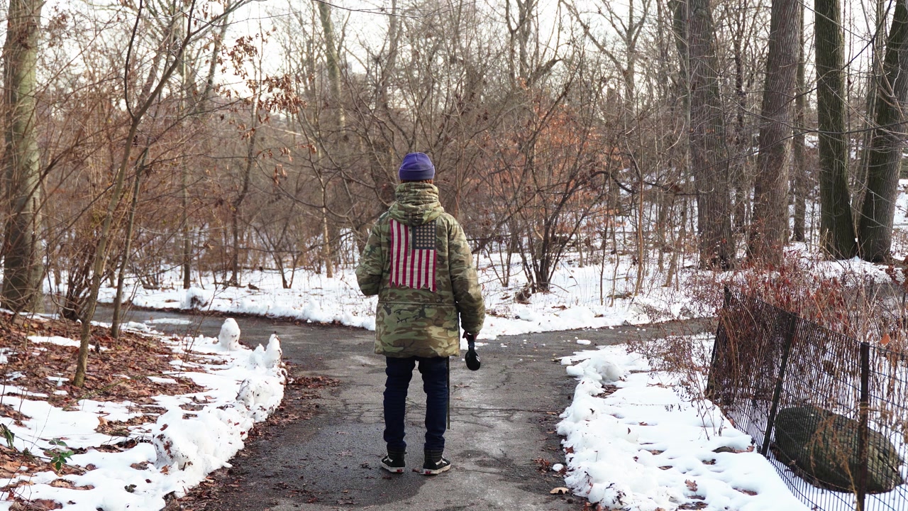 Un giovane uomo, vestito con un cappotto mimetico con una bandiera americana, si trova a un bivio in un paesaggio invernale innevato. Tiene delle cuffie nella mano destra, perso nei pensieri, contempla il suo prossimo passo