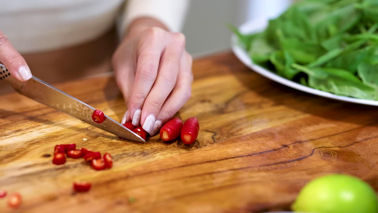 Hands slicing red chilies on a wooden board with fresh greens nearby, under bright lighting