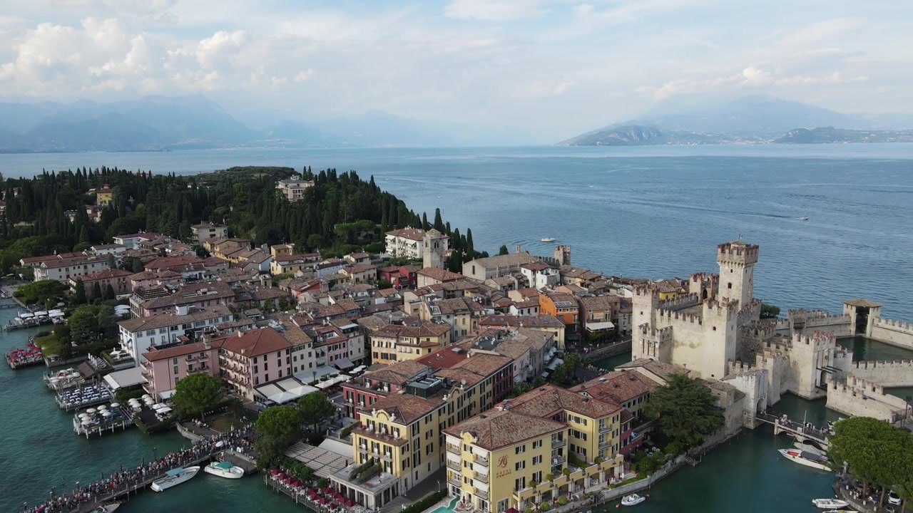 Riprese aeree spettacolari del Castello Scaligero medievale di Sirmione e degli edifici color pastello in riva al lago, circondati dal blu del Lago di Garda, da una vegetazione lussureggiante e da nuvole soffici, sotto una luce diurna cinematografica.