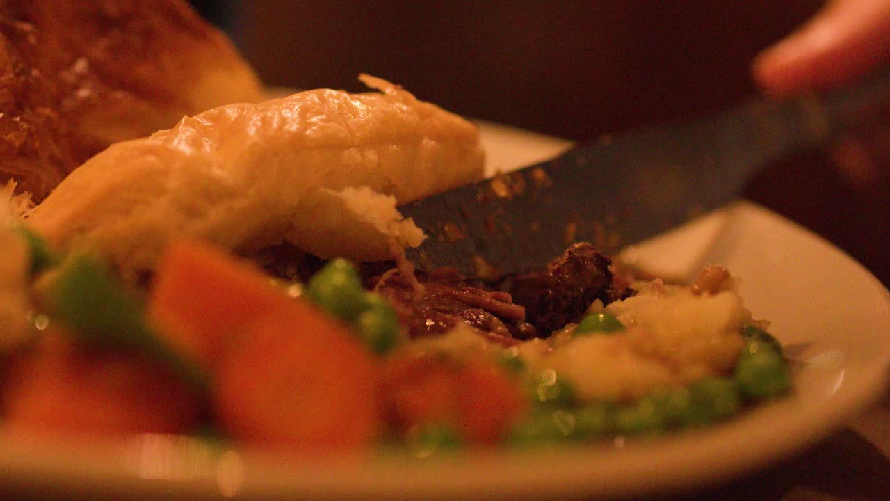 Hand slices steak pie, revealing filling, with peas, carrots, and mashed potatoes under warm lighting