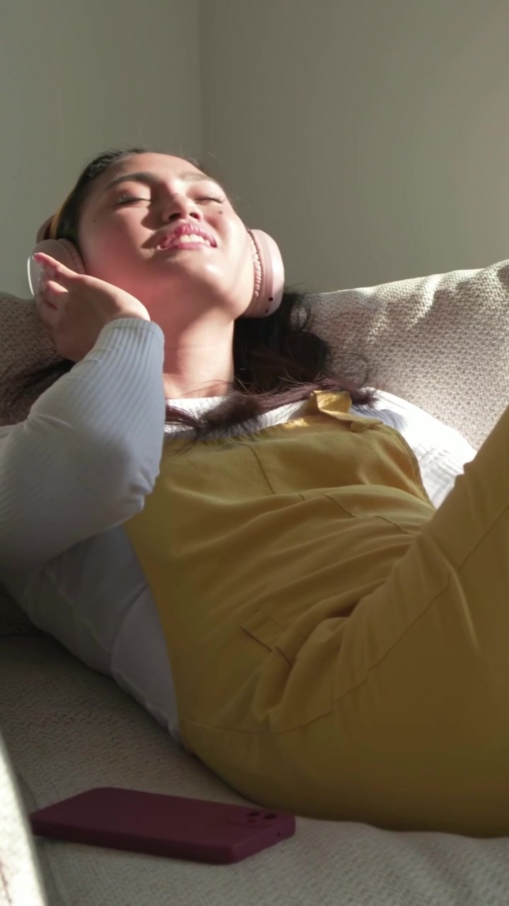 Video verticale: Donna sdraiata in color senape che sistema le cuffie rosa sul divano, il sole che attira lo sguardo, controlla il cellulare