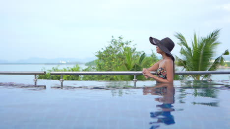 while leaning on the rail on the edge of an infinity pool, a young woman in a floppy sun hat and bathing suit looks from the ocean in the background to a point on her left