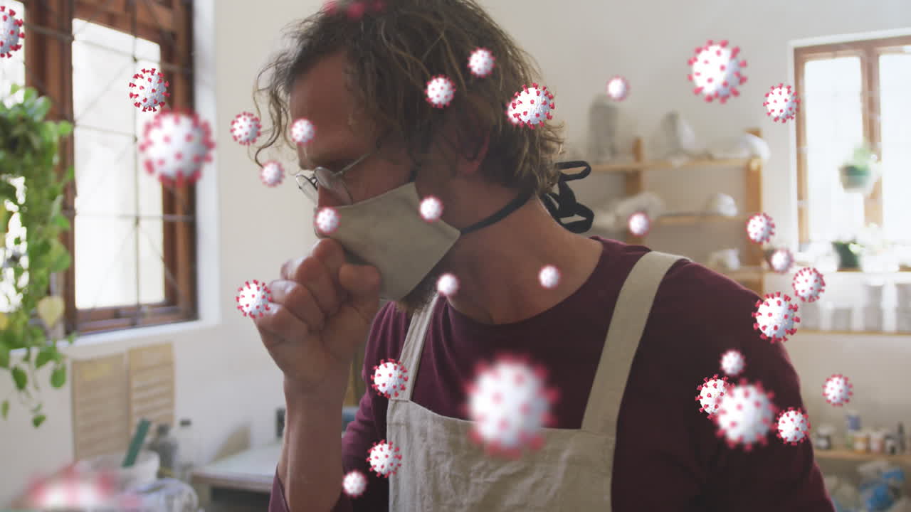 코로나바이러스 세포의 애니메이션이 표면 마스크를 쓴 백인 남자가 도자기 작업장에서 기침을 하고 있다.