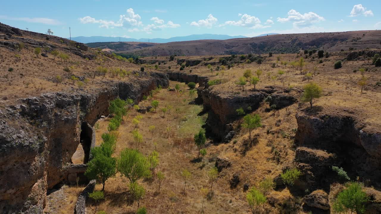 vuelo inverso en un pequeño cañón de piedra caliza con árboles y vemos una estructura de piedra utilizada como un recinto para los animales hay hierba seca en una mañana de verano con un cielo azul segovia españa