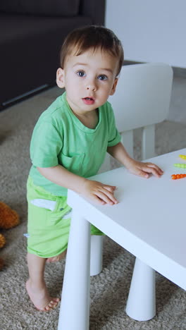 pretty little toddler is in his room. child wants to sit at the table with a range of bright toys on. view from top. vertical video