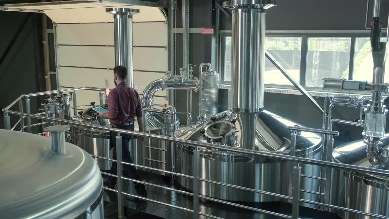 Young male brewer wearing a leather apron supervise the process of beer fermentation at a modern brewery factory
