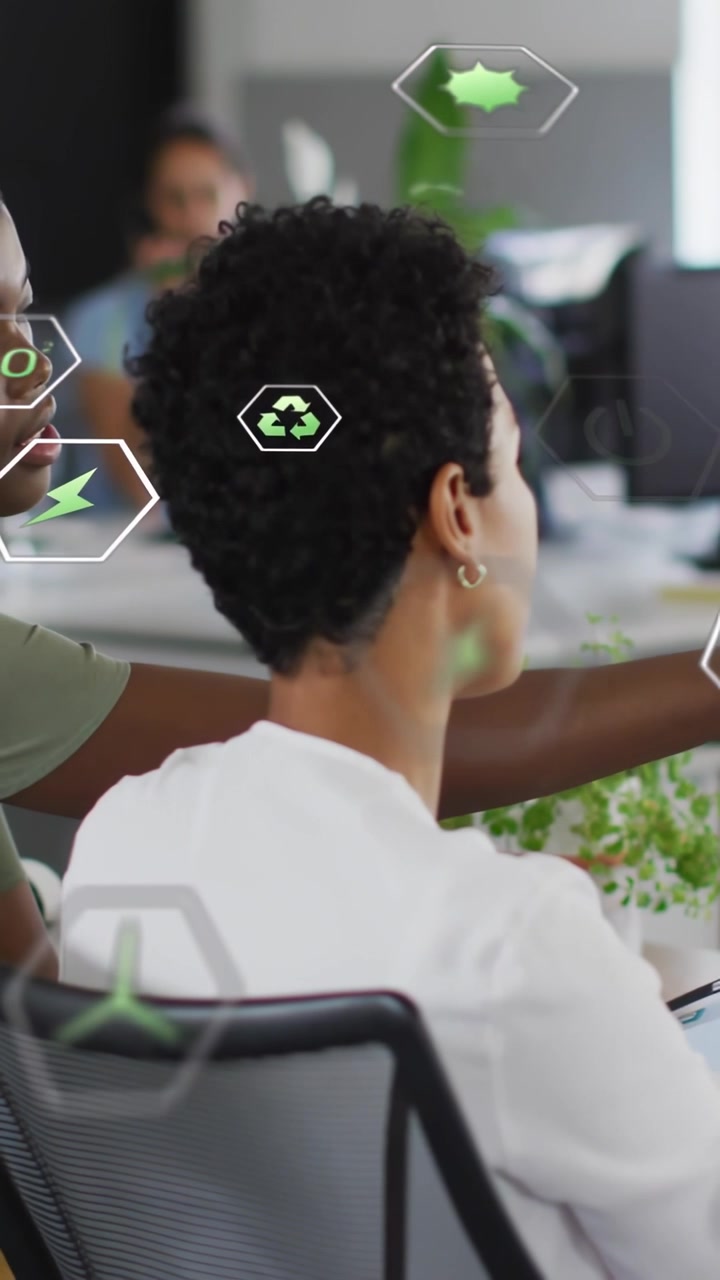 Vertical video: Seated woman reaching monitor as colleague leaning in, guiding eco holograms for UX