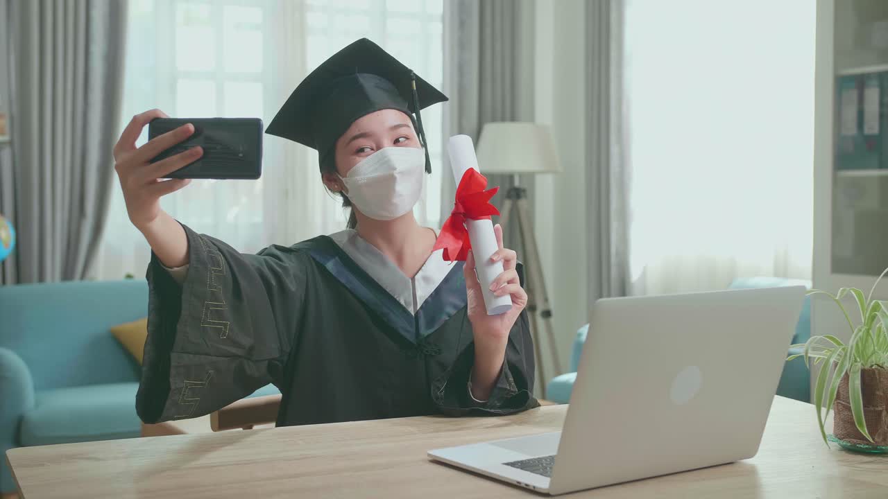 Young Asian Woman Wearing Protection Face Mask, Taking A Selfie While Wearing A Graduation Gown And Cap, Hand Holding University Certificate In Living Room