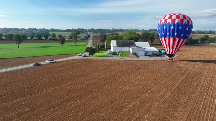 Drone approaching shot of american hot air balloon on farm field near farm house and driving cars on road