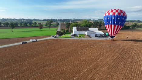 Drone-approaching-shot-of-american-hot-air-balloon-on-farm-field-near-farm-house-and-driving-cars-on-road