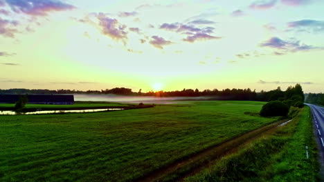 Aerial-drone-forward-moving-shot-over-pond-beside-green-grasslands-surrounded-by-trees-on-a-foggy-autumn-sunrise-beside-a-highway