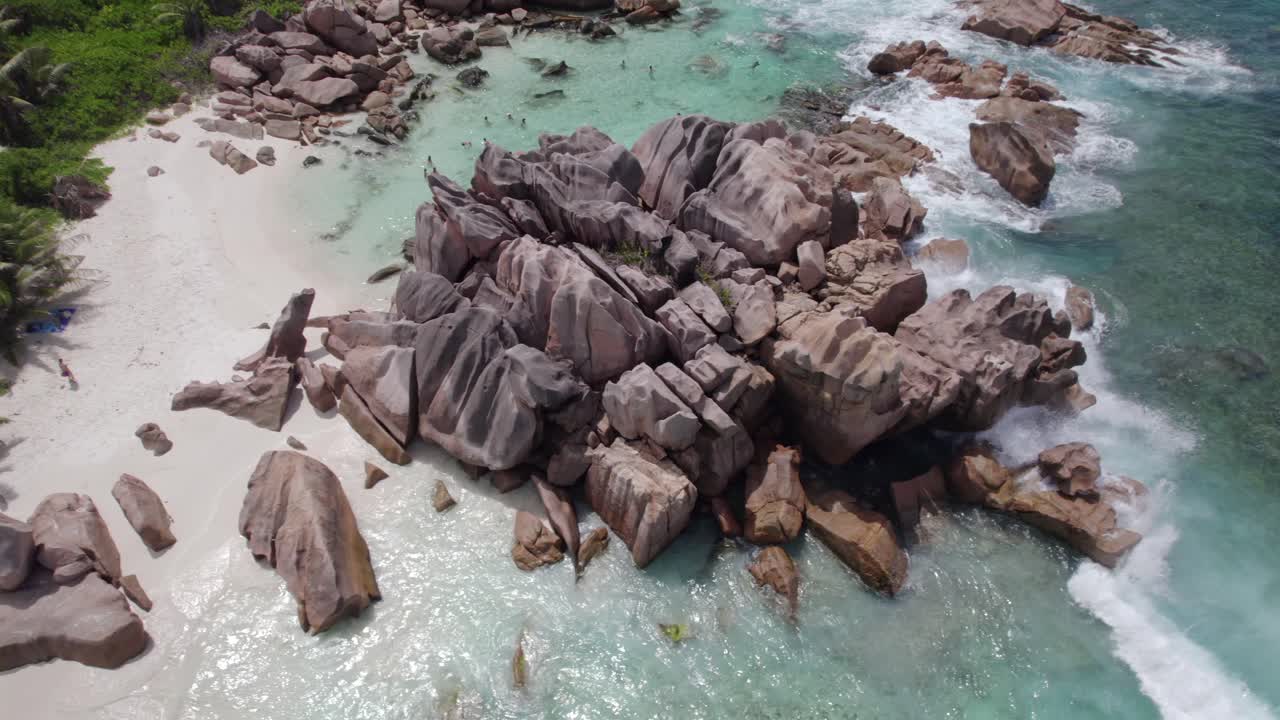 Pemandangan drone mata burung yang diambil dari ketinggian, lalu menggeser ke bawah, memperlihatkan batu-batu granit dekat garis pantai, air biru kehijauan yang jernih, orang-orang yang sedang snorkeling, pantai berpasir putih, Anse Cocos, La Digue, Seychelles 010.