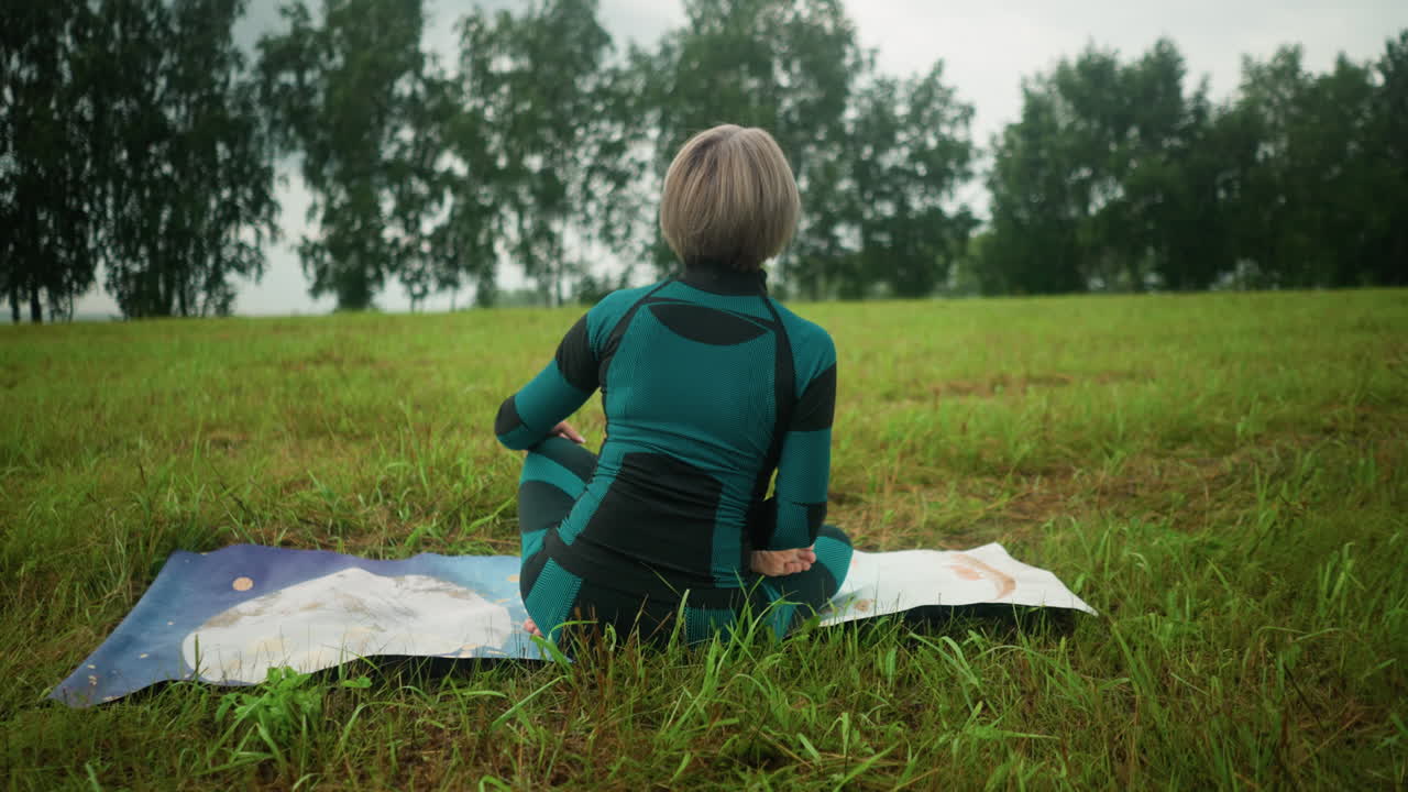 시다사나 포즈에서 다채로운 매트에 앉아있는 노인 여성의 뒷면, 구름이 가득한 하늘 아래 광활한 잔디에서 요가를 연습하고, 배경에는 나무가 줄지어 있습니다.