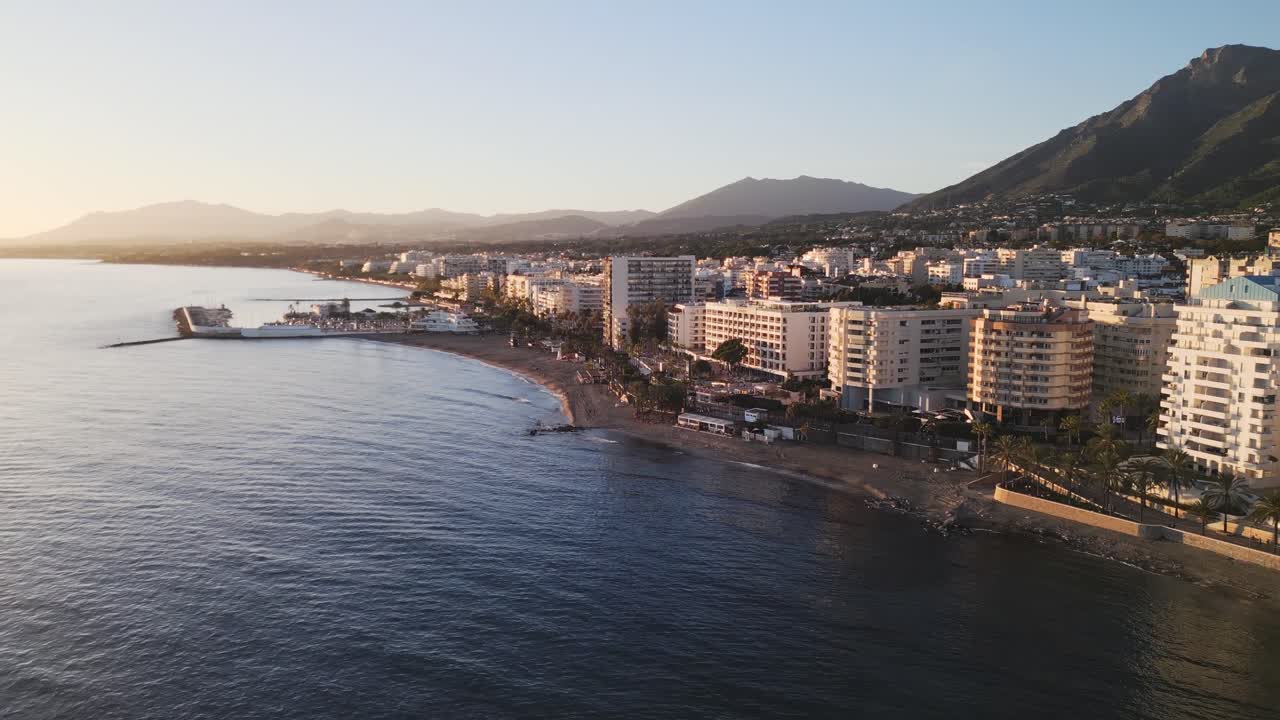 Panoramica aerea verso destra lungo il lungomare di Marbella durante l'ora d'oro, edifici di appartamenti costieri di lusso e hotel con vista sulle calme acque del Mediterraneo, Spagna meridionale