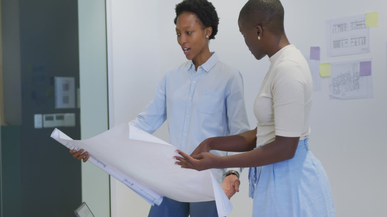 Two happy african american female architects in discussion studying blueprints in office
