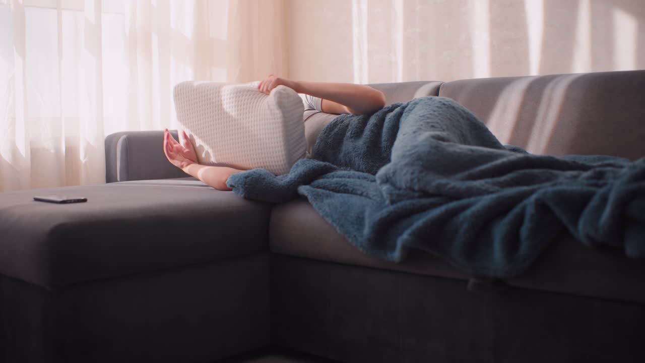 Sleepy girl curled on couch holding pillow close while sunlight filters through curtain, covering part of face with arm, wrapped in soft blue blanket, resisting morning light in quiet interior