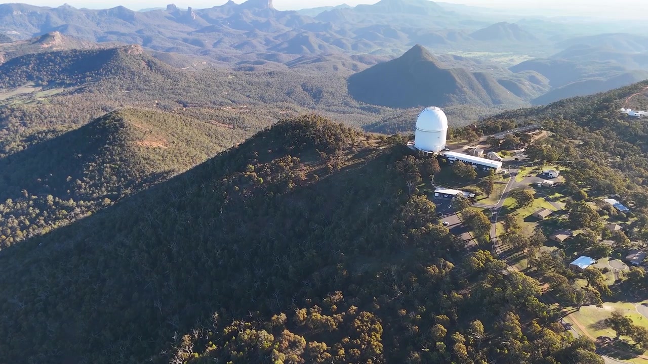 Bredt luftfoto av teleskopkuppelen ved Siding Spring Observatory på toppen av en fjellkjede.