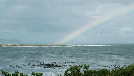 Seagulls-flying-over-coastline-with-rainbow-over-ocean-in-distance,-Overstrand