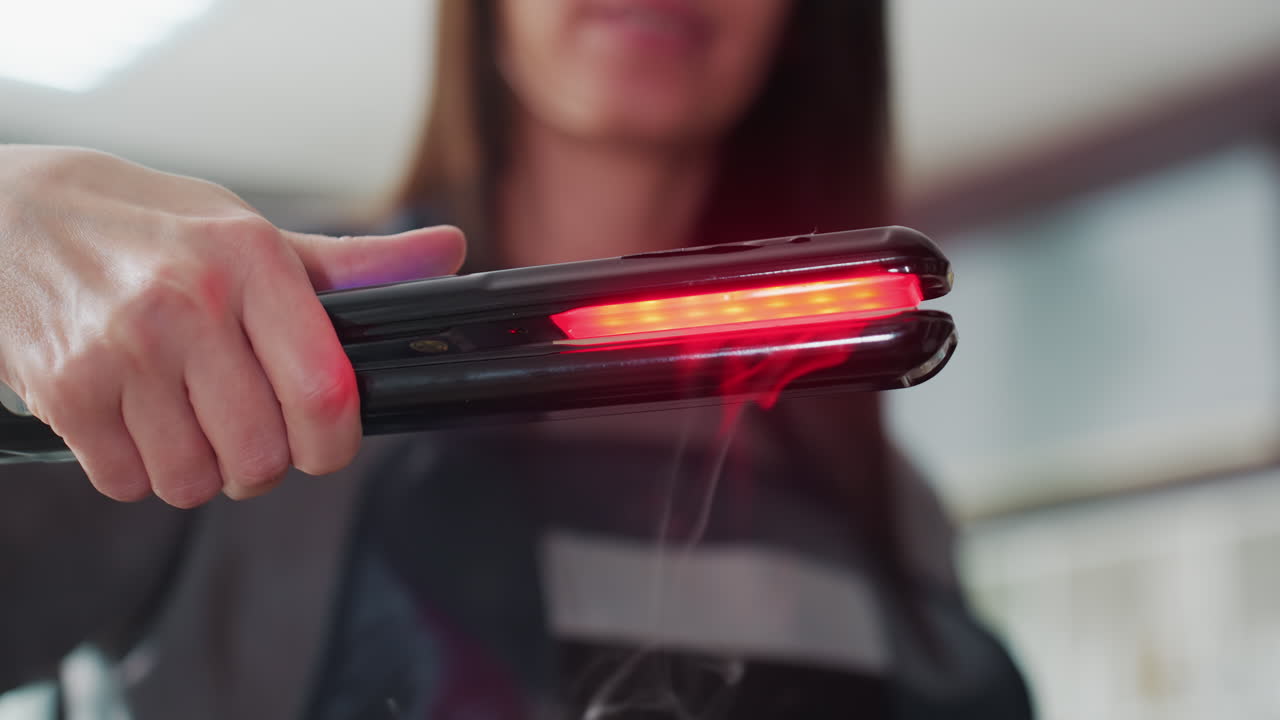 Estilista sosteniendo una plancha iluminada con luz roja cerca de la cámara; persona caucásica enmarcada por el interior de un salón; toma de presentación del producto con placas brillantes y una presentación segura; ambiente publicitario