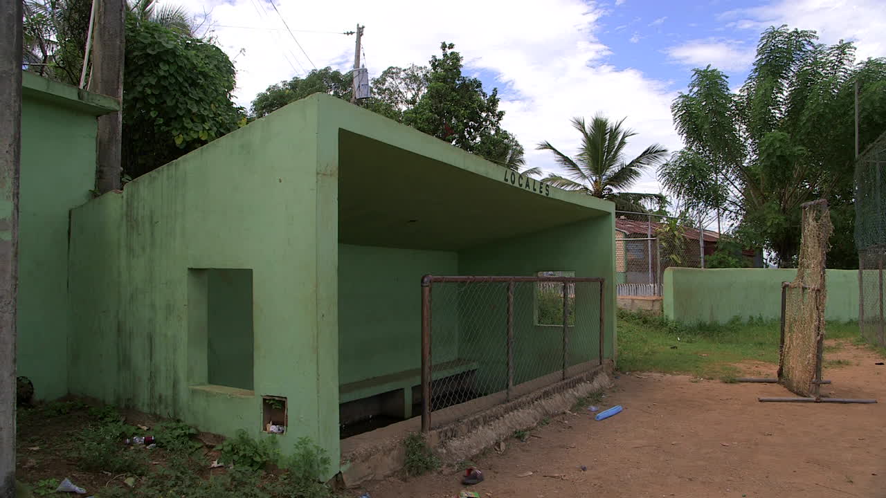 Dugout verde abbandonato su un campo da baseball polveroso nella Repubblica Dominicana rurale, con vernice scrostata, rete metallica, erba incolta e senza giocatori sotto un cielo tropicale nuvoloso.
