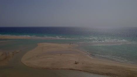 Sotavento-Beach,-Fuerteventura:-wonderful-aerial-view-traveling-in-the-fantastic-beach-with-people-enjoying-it,-on-a-sunny-and-hazy-day