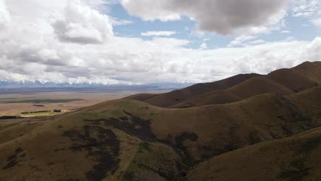 Aerial---brown,-dry-ridge-line-in-New-Zealand,-vast-plains-and-snow-capped-mountains-in-background