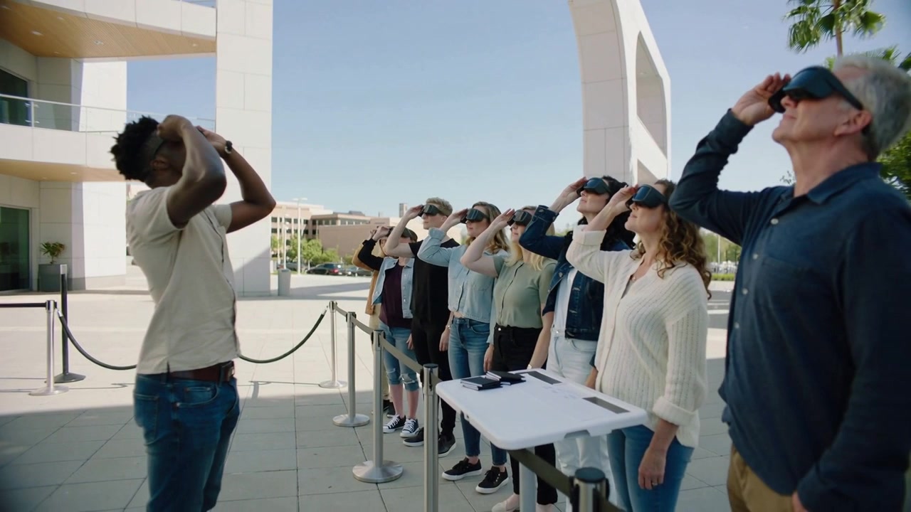 Guida che alza l'auricolare, indossa una maglietta a maniche corte e jeans, e guida la fila nella piazza per una demo VR, con spazio per il testo.