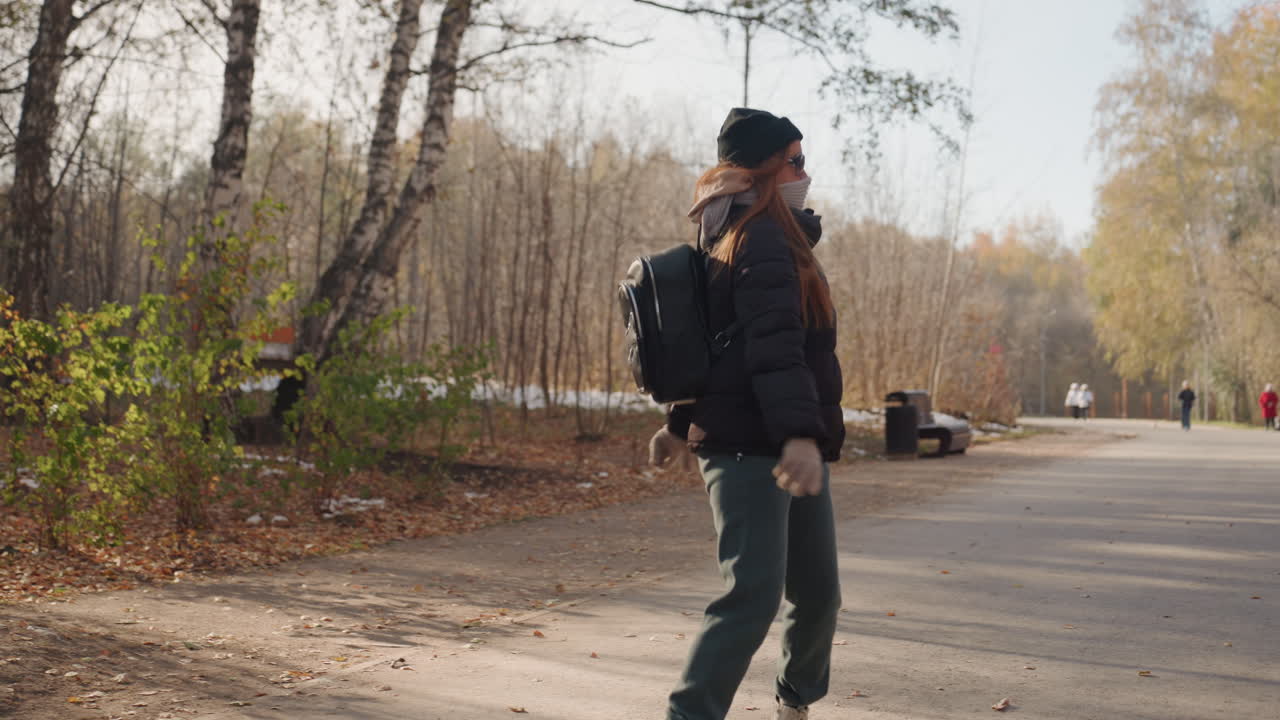 Female walking near wooded park edge, glancing sideways while pacing along paved path, wearing warm jacket, beanie and backpack with face mask, scattered leaves and soft sunlight, calm vigilant mood