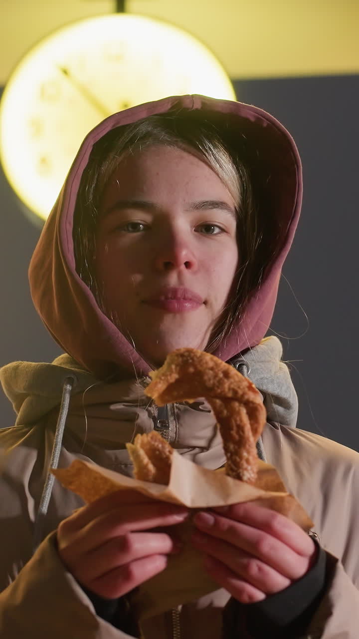 mujer joven sonriente de pie al aire libre por la noche sosteniendo pastel crujiente en la mano, brillo cálido del reloj de la calle iluminado en el fondo