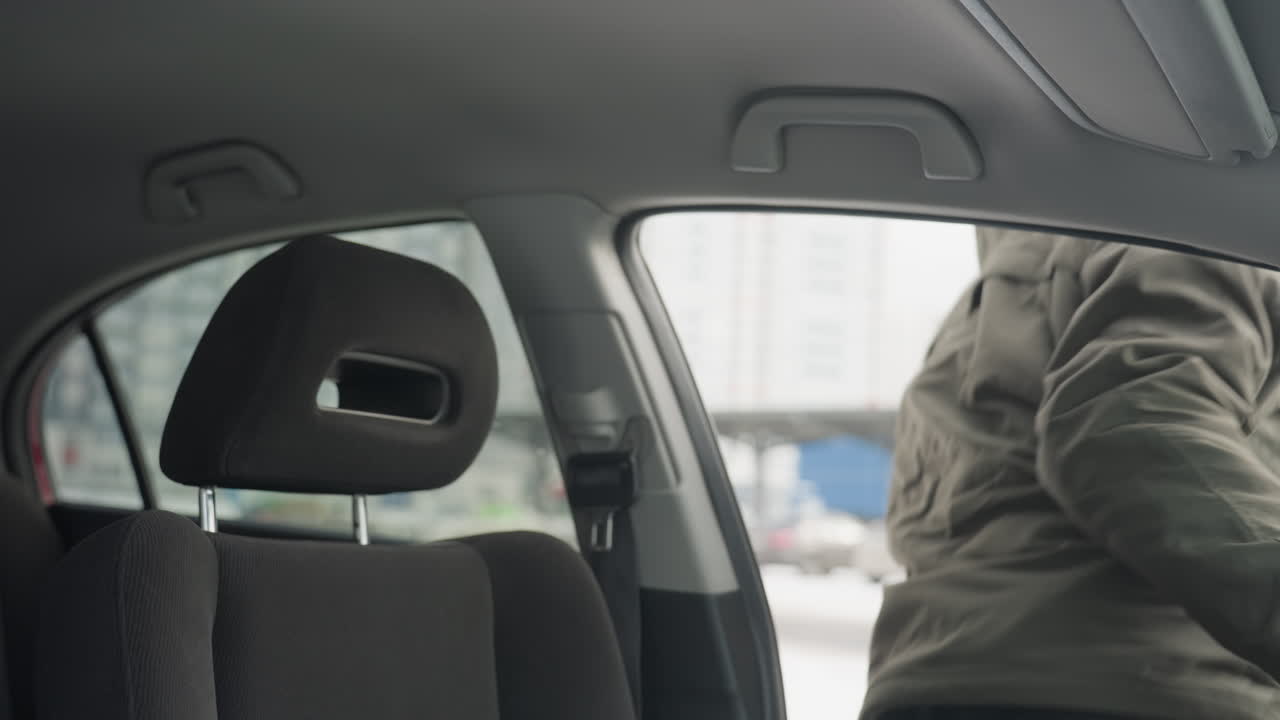 close up of man in glasses and green winter jacket stepping out of car, snowy urban environment visible through window with parked cars and building blurred in background