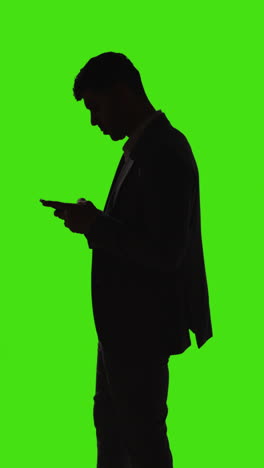 Vertical-Video-Silhouette-Of-Businessman-Standing-Looking-At-Screen-Of-Mobile-Phone-Against-Green-Screen-Background