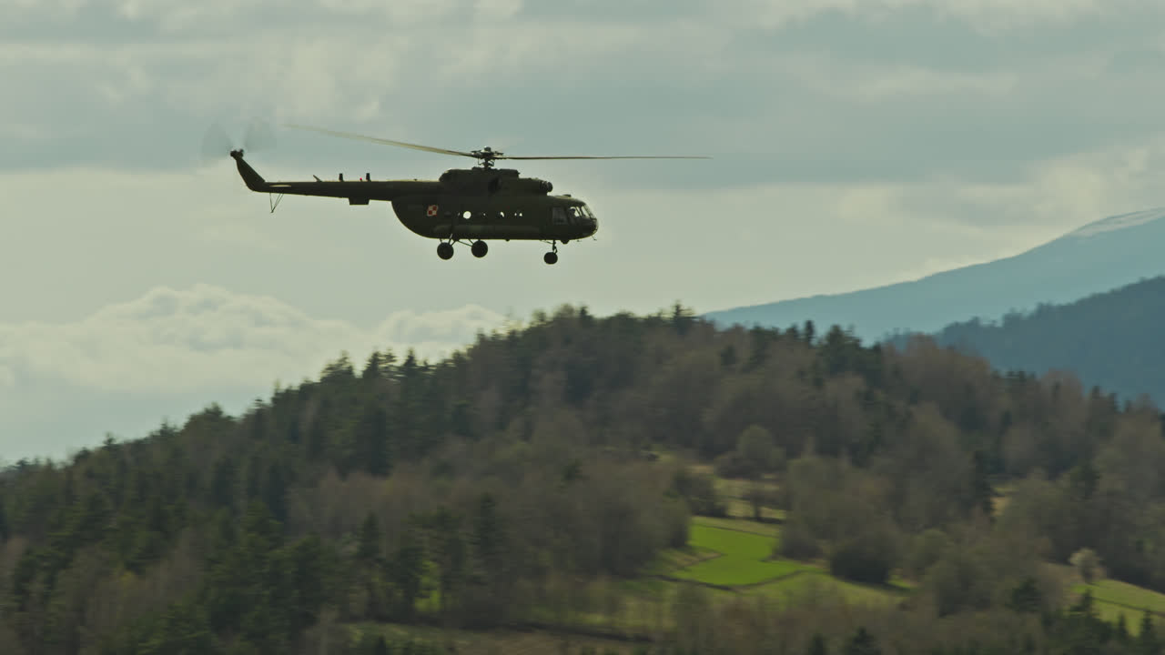 Silhouette of a helicopter in flight captured against a pale sky and distant rolling mountains. The image conveys a sense of observation and remote patrol in a serene, highland environment.