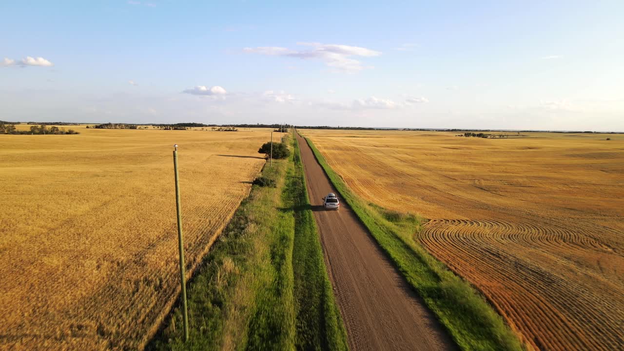 Hopeinen auto ajaa pitkää, sähkepylväiden reunustamaa hiekatietä Amerikan maaseudulla auringonlaskun aikaan.