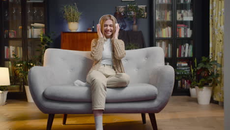 happy young woman listening music in wireless headphones while sitting on a couch at home