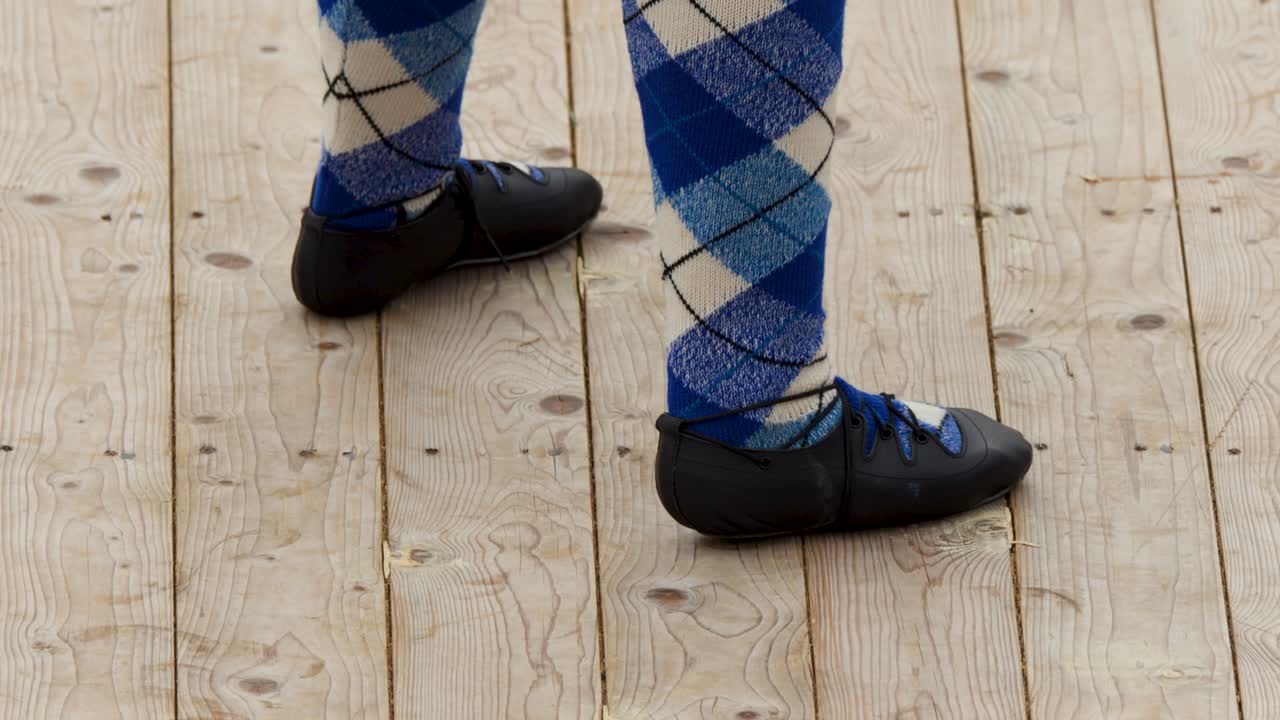 Dancer in argyle socks performs Highland dance steps on wooden platform, natural daylight, static camera