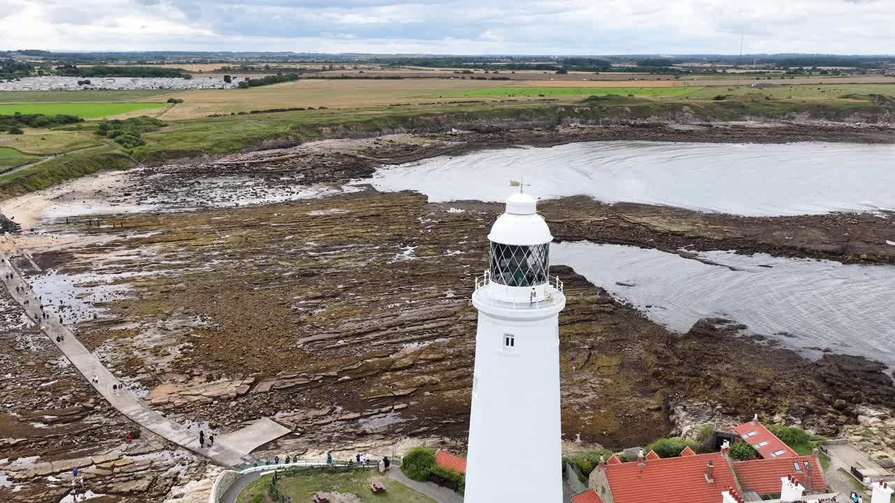 Drone camera smoothly circles a tall white lighthouse on a rugged coastline, revealing tidal flats, nearby buildings, and overcast daylight conditions