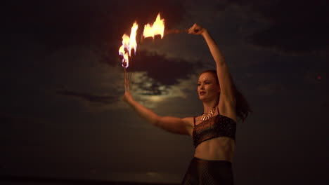 woman dancing with fire fans against cloudy full moon night sky, medium shot slowmo