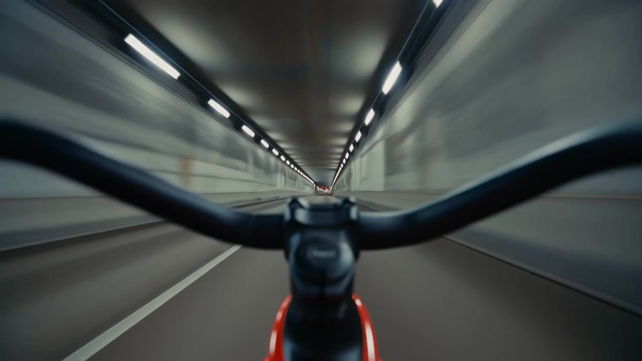 Low rider view moves through illuminated road tunnel with bicycle handlebars framing foreground, repeating ceiling lights, glossy metal surfaces, and converging lines create speed, depth, and urban
