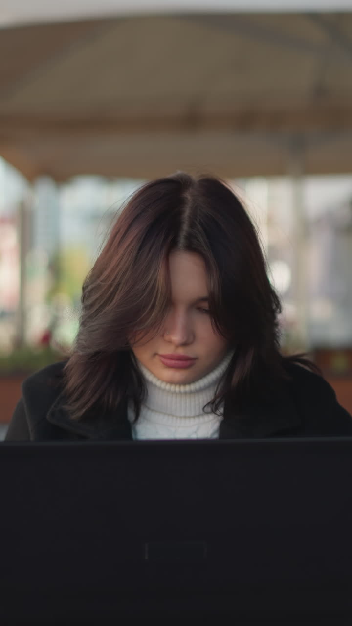 freelancer centrado en la pantalla de la computadora portátil sentado al aire libre en un entorno urbano con luz natural, muebles modernos, fondo borroso de edificios, flores y sombrillas de patio revoloteando suavemente