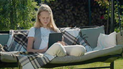 a teenage girl uses a tablet, sits on a garden swing in the backyard of a house.