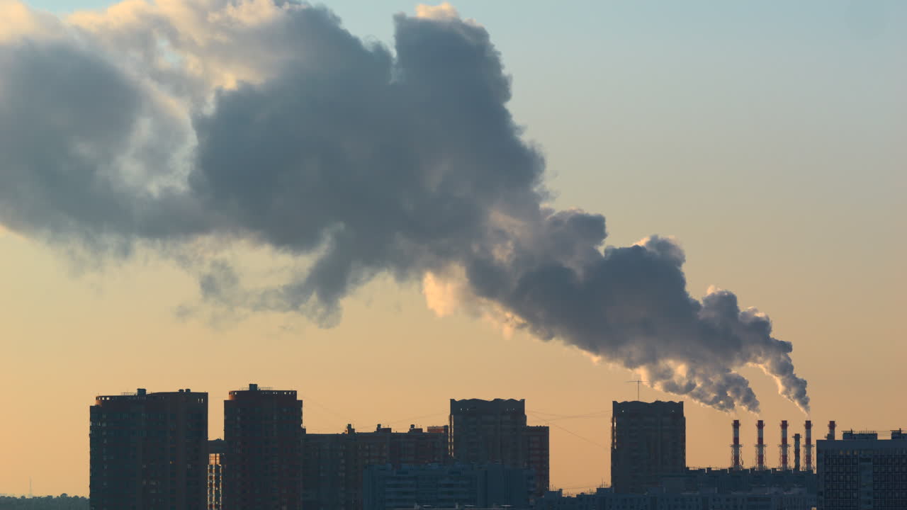 Multiple industrial chimneys releasing a huge plume above residential buildings in warm evening light Urban skyline with heavy smoke steam haze and atmospheric backlit pollution concept city life