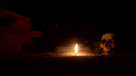 wizard's desk. a desk lit by candle light. a human skull, old books on sand surface. halloween still-life background with a different elements on dark toned foggy background. slider shot